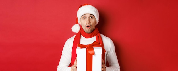 Concept de vacances d'hiver, Noël et mode de vie. Gros plan sur un beau mec surpris en bonnet et écharpe, l'air étonné et tenant un cadeau du nouvel an, debout sur fond rouge.