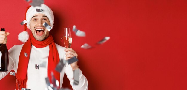 Concept de vacances d'hiver, de noël et de style de vie. Gros plan sur un homme heureux célébrant le nouvel an, tenant une bouteille de champagne et un verre, debout sur fond rouge avec des confettis