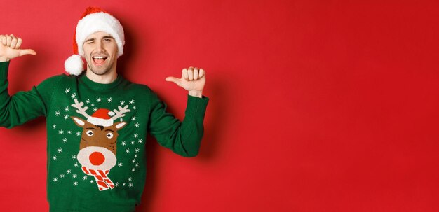 Concept de vacances d'hiver noël et style de vie Sassy bel homme en bonnet de noel et pull vert pointant vers lui-même et clignant de l'oeil debout sur fond rouge