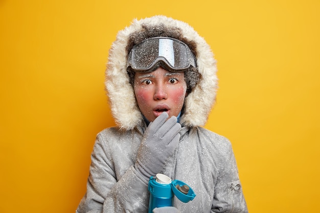 Concept de vacances d'hiver. Surpris femme active ethnique apprécie l'activité sportive garde la bouche ouverte de boissons choc boisson chaude vêtue de vêtements d'extérieur.