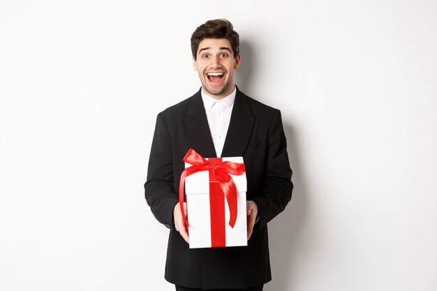 Concept de vacances de Noël, de célébration et de style de vie. Image d'un beau mec en costume noir qui a l'air excité, a un cadeau, debout sur fond blanc