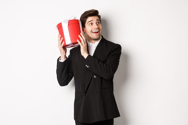 Concept de vacances de Noël, de célébration et de style de vie. Image d'un homme séduisant en costume noir, secouant le présent pour deviner quoi à l'intérieur, profitant du nouvel an, debout sur fond blanc.