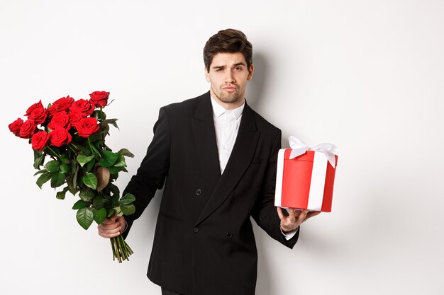 Concept de vacances, de relation et de célébration. Bel homme confiant en costume noir, allant à un rendez-vous, tenant un bouquet de roses et présent, debout sur fond blanc