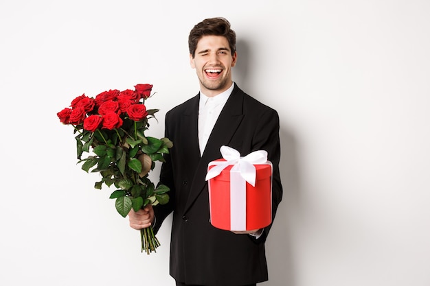 Concept de vacances, de relation et de célébration. Charmant jeune homme en costume noir, tenant une boîte-cadeau et un bouquet de roses, un clin d'œil et souriant, debout sur fond blanc
