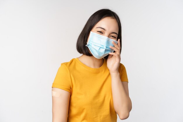 Concept de vaccination et de soins de santé Covid19 Jolie fille asiatique dans un masque médical montrant un pansement après la vaccination contre le coronavirus debout sur fond blanc