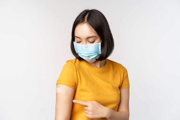 Concept de vaccination et de soins de santé Covid19 Jolie fille asiatique dans un masque médical montrant un pansement après la vaccination contre le coronavirus debout sur fond blanc