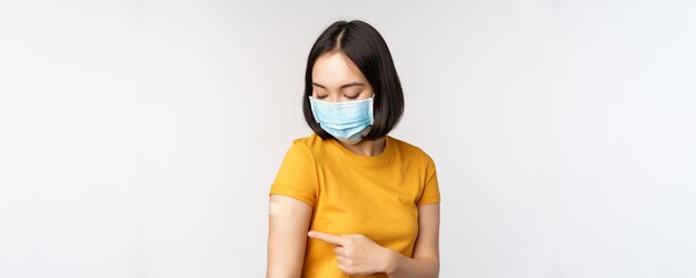 Concept de vaccination et de soins de santé Covid19 Jolie fille asiatique dans un masque médical montrant un pansement après la vaccination contre le coronavirus debout sur fond blanc