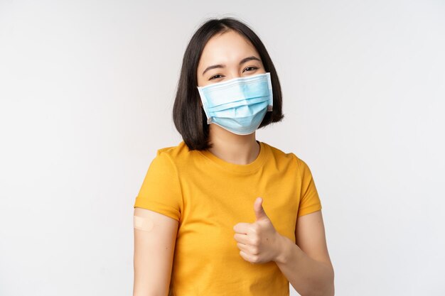 Concept de vaccination et de soins de santé Covid19 Portrait d'une jolie fille asiatique portant un masque médical a un pansement sur l'épaule après le vaccin contre le coronavirus debout sur fond blanc