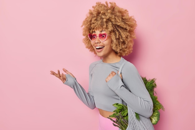 Photo gratuite concept végétarien une femme surprise positive porte un sac en tissu avec des légumes frais achetés sur le marché mange des vitamines printanières porte des lunettes de soleil à la mode top court regarde joyeusement de côté se tient à l'intérieur