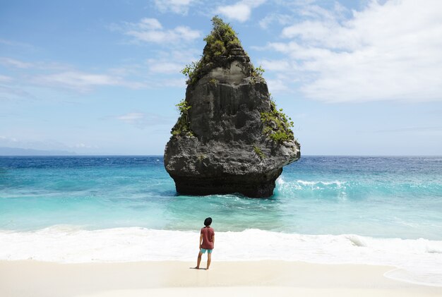 Concept de voyage, aventure, passe-temps et vacances. Jeune homme habillé de façon décontractée au chapeau noir marchant le long de la plage de sable déserte, face à l'océan turquoise avec île de pierre avec de hauts rochers au milieu