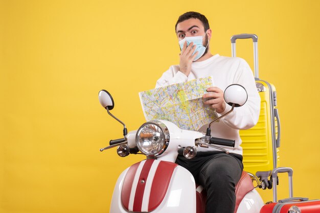 Concept de voyage avec un gars surpris en masque médical assis sur une moto avec une valise jaune dessus et tenant une carte en jaune