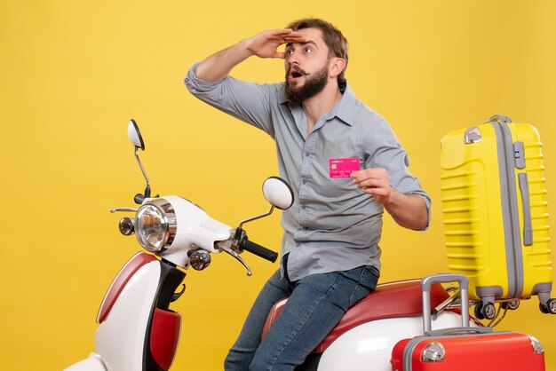 Concept de voyage avec jeune homme barbu concentré émotionnel assis sur une moto et tenant une carte bancaire dessus sur jaune