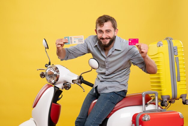 Concept de voyage avec jeune homme barbu confiant assis sur une moto tenant une carte bancaire et un billet sur elle sur jaune