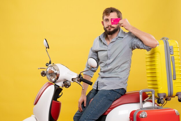 Concept de voyage avec jeune homme barbu confiant assis sur une moto tenant une carte bancaire sur elle sur jaune