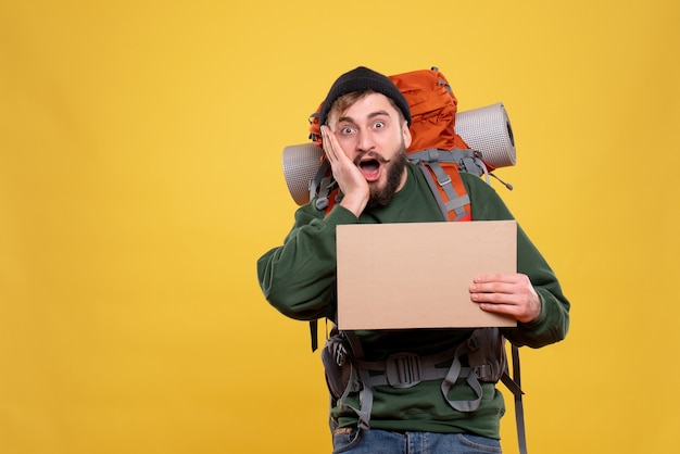 Concept de voyage avec jeune homme surpris avec packpack et tenant une feuille sans écrire dessus sur jaune