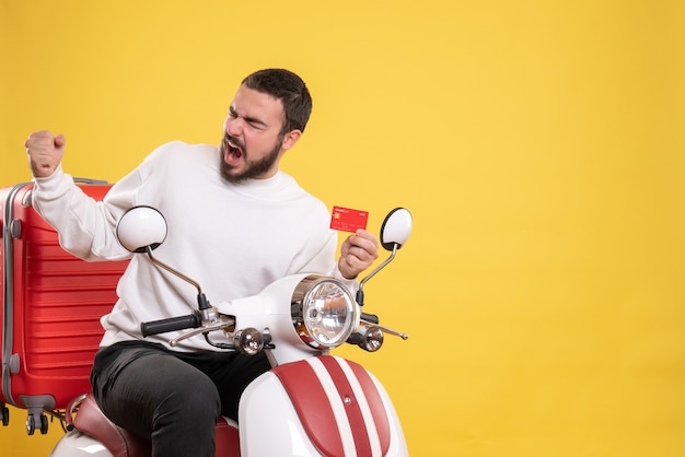 Photo gratuite concept de voyage avec un jeune homme de voyage ambitieux et fier assis sur une moto avec une valise dessus tenant une carte bancaire sur jaune