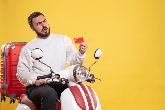 Concept de voyage avec un jeune homme de voyage confus assis sur une moto avec une valise dessus tenant une carte bancaire sur jaune