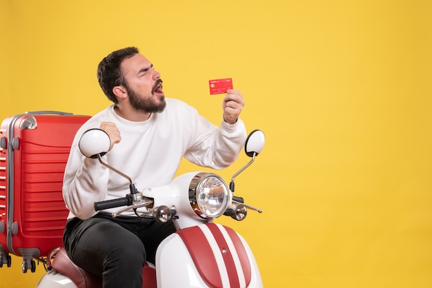 Concept de voyage avec un jeune homme de voyage heureux et émotif assis sur une moto avec une valise dessus montrant une carte bancaire en jaune