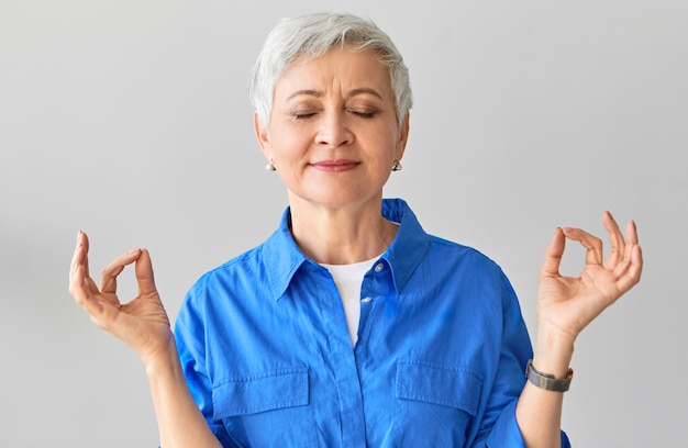 Concept zen, sagesse, équilibre et relaxation. Belle femme aux cheveux gris dans la cinquantaine posant avec les yeux fermés méditant après le yoga reliant le pouce et l'index en geste mudra