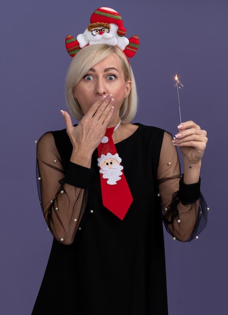 Photo gratuite concerné femme blonde d'âge moyen portant bandeau et cravate du père noël tenant vacances sparkler gardant la main sur la bouche isolé sur mur violet