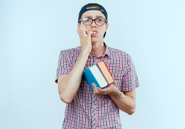 concerné jeune étudiant garçon portant un sac à dos et des lunettes et une casquette tenant des livres et se mord les ongles