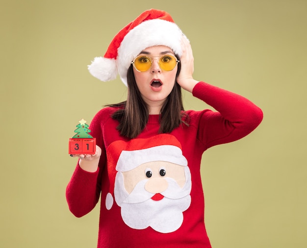 Concerné jeune jolie fille caucasienne portant pull du père noël et bandeau avec des lunettes tenant jouet arbre de Noël avec date