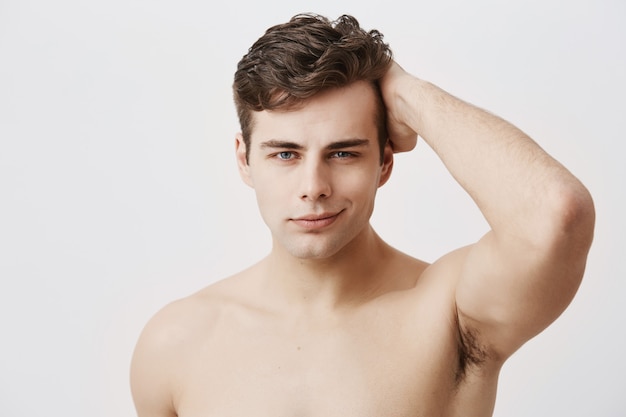 Condifent jeune homme européen avec une coupe élégante et des yeux attrayants, étant nu, touchant les cheveux noirs, posant. Beau modèle masculin avec une peau propre et saine, souriant doucement