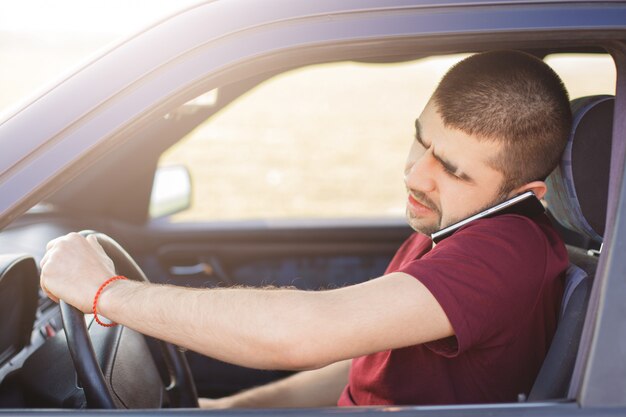 Un conducteur masculin au look concentré conduit la voiture et parle sur téléphone mobile car il résout des problèmes importants à distance, voyage sur de longues distances