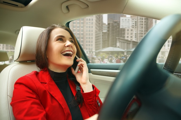 Conduire En Ville. Jeune Femme Séduisante Au Volant D'une Voiture. Jeune Modèle Assez Caucasien En Veste Rouge élégante Et élégante Assis à L'intérieur Du Véhicule Moderne