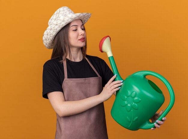 Confiant assez caucasien femme jardinier portant chapeau de jardinage tenant et regardant arrosoir sur orange