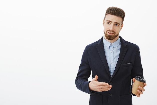 Confiant beau homme d'affaires européen avec barbe et moustache en costume élégant faisant des gestes discutant des affaires tenant une tasse de café traitant avec des partenaires de l'entreprise contre le mur blanc