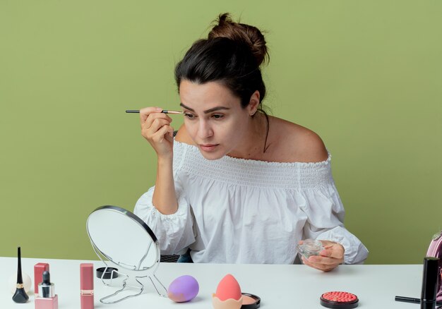Confiant belle fille est assise à table avec des outils de maquillage regarde miroir appliquant le fard à paupières avec pinceau de maquillage isolé sur mur vert