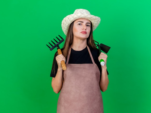 Photo gratuite confiant belle fille de jardinier en uniforme portant chapeau de jardinage mettant râteau avec houe râteau sur l'épaule isolé sur vert