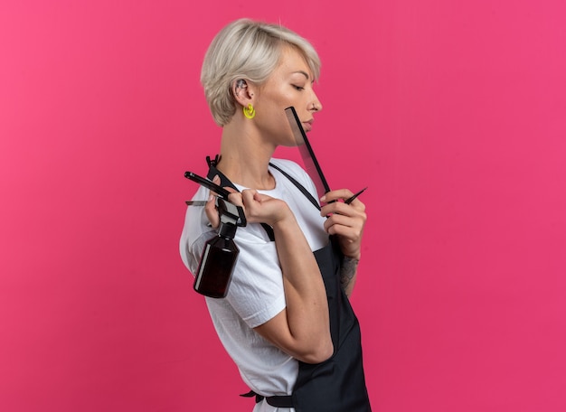 Confiant debout dans la vue de profil jeune belle femme barbier en uniforme tenant des outils de barbier isolés sur un mur rose