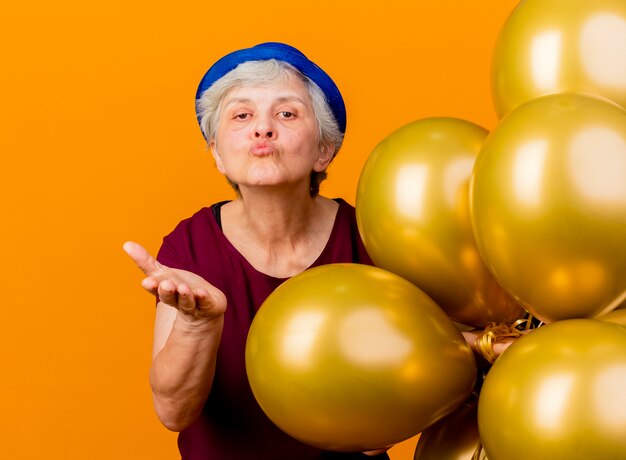 Confiant femme âgée portant chapeau de fête détient des ballons d'hélium et envoie un baiser avec la main isolée sur le mur orange