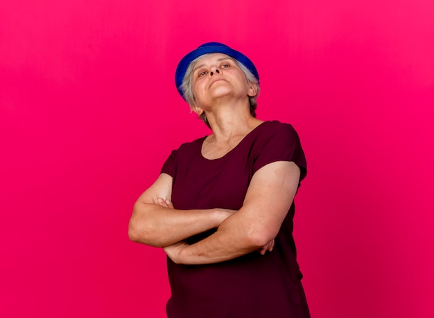 Confiant femme âgée portant chapeau de fête se tient avec les bras croisés sur rose