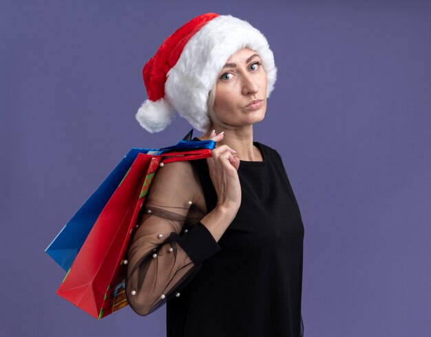 Confiant femme blonde d'âge moyen portant un chapeau de Noël debout en vue de profil tenant des sacs-cadeaux de Noël sur l'épaule en regardant la caméra isolée sur fond violet avec copie espace