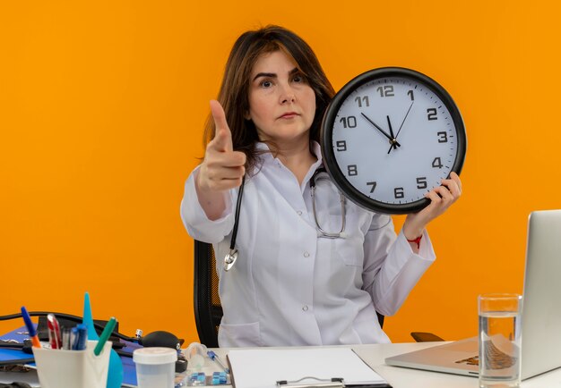 Confiant femme médecin d'âge moyen portant une robe médicale et un stéthoscope assis au bureau avec presse-papiers d'outils médicaux et ordinateur portable tenant horloge pointant isolé