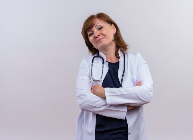 Confiant femme médecin d'âge moyen portant une robe médicale et stéthoscope debout avec une posture fermée sur un mur blanc isolé avec copie espace