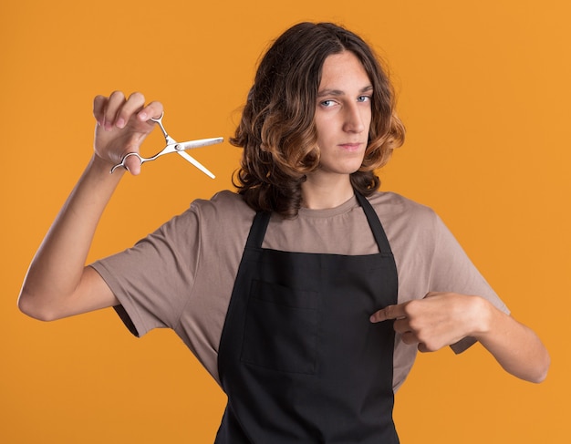 Confiant jeune beau barbier en uniforme tenant des ciseaux pointant sur lui-même