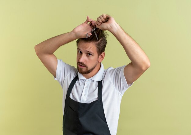 Confiant jeune beau coiffeur couper ses cheveux avec des ciseaux isolé sur vert olive avec copie espace