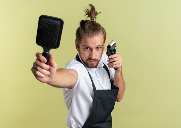 Confiant jeune beau coiffeur étirant le peigne et tenant une tondeuse à cheveux isolé sur vert olive avec espace copie