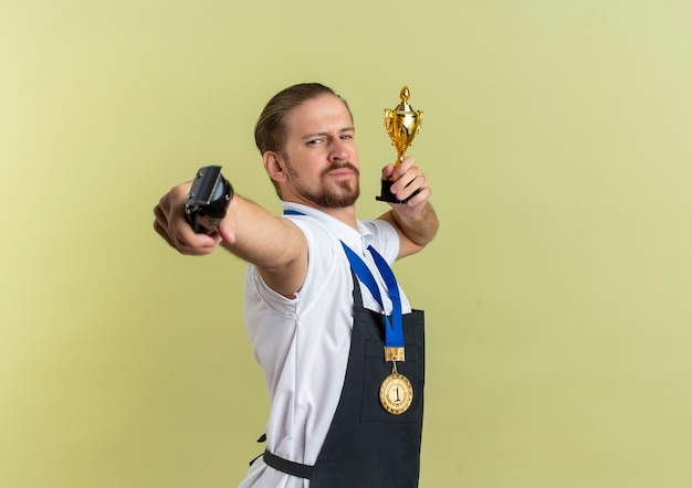 Confiant jeune beau coiffeur portant la médaille autour du cou étirant les tondeuses à cheveux et tenant la coupe du gagnant isolé sur vert olive avec espace copie