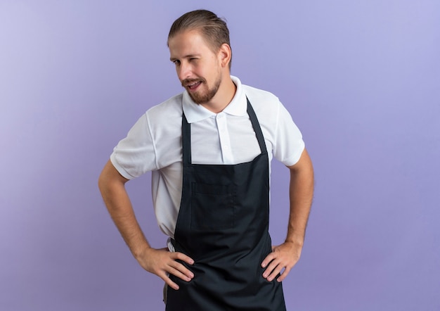 Confiant jeune beau coiffeur portant un uniforme clignant des yeux et mettant les mains sur la taille isolé sur violet avec copie espace