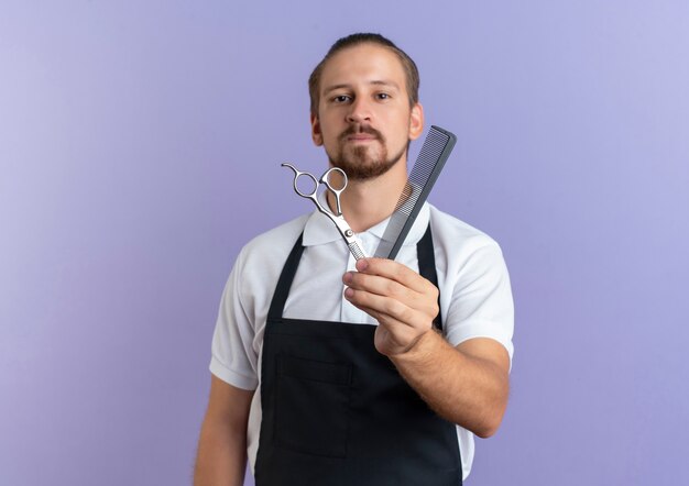 Confiant jeune beau coiffeur portant l'uniforme s'étendant sur le peigne et les ciseaux vers la caméra isolée sur violet avec copie espace