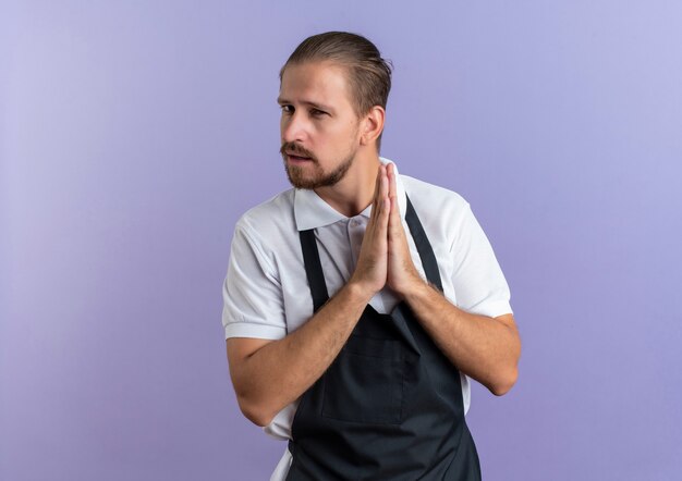 Confiant jeune beau coiffeur en uniforme gardant les mains ensemble isolé sur violet avec copie espace