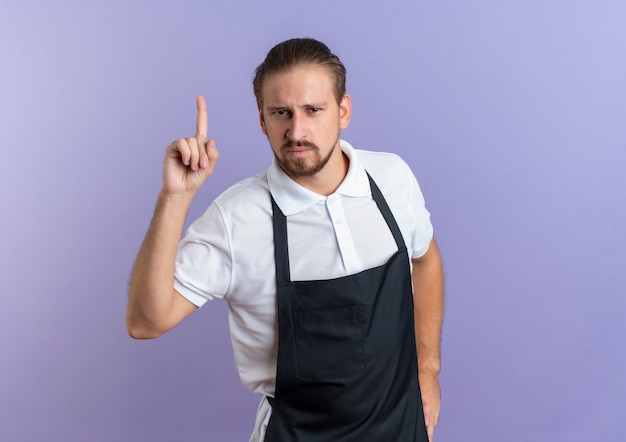 Photo gratuite confiant jeune beau coiffeur en uniforme mettant la main sur la taille et pointant vers le haut isolé sur violet avec copie espace