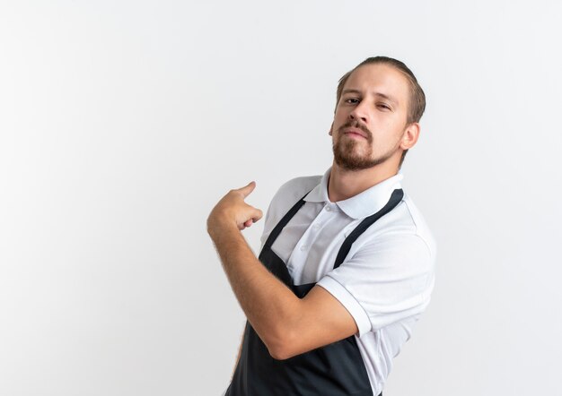 Confiant jeune beau coiffeur en uniforme pointant derrière isolé sur blanc avec copie espace