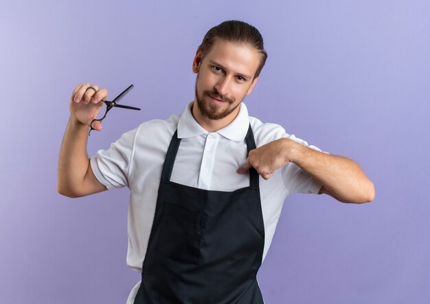 Confiant jeune beau coiffeur en uniforme tenant des ciseaux et pointant sur lui-même isolé sur violet