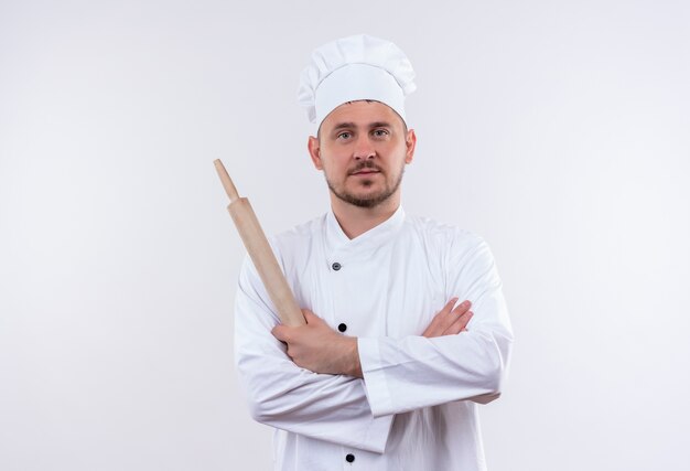 Confiant jeune beau cuisinier en uniforme de chef debout avec une posture fermée et tenant un rouleau à pâtisserie isolé sur un mur blanc